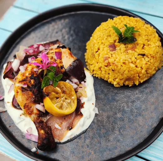 Greek-style chicken over tzatziki with charred lemon and red onion, beside a dome of yellow rice