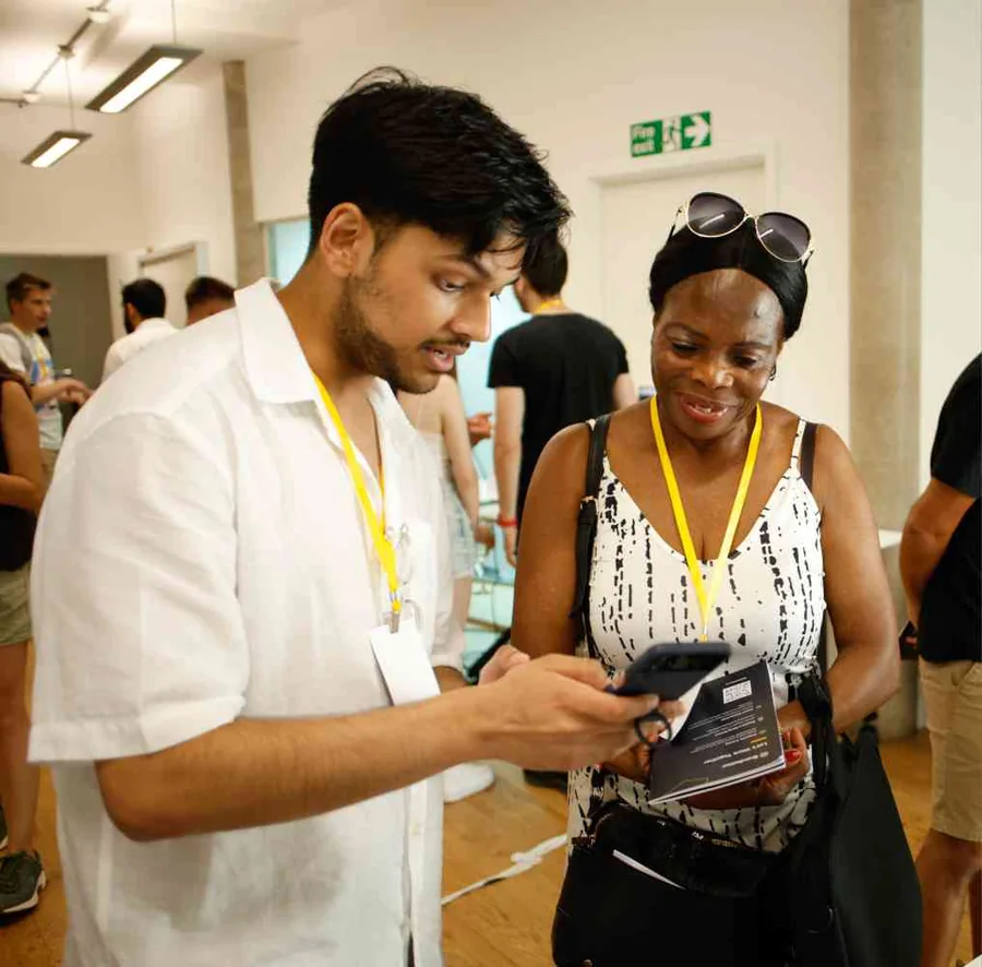 Mahin showing his project on his phone to a visitor at the UX bootcamp&rsquo;s Demo Day, both wearing yellow lanyards